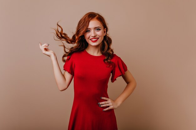 笑っている赤いドレスを着たスリムな白人女性。写真撮影中に髪の毛で遊ぶゴージャスな生姜の女の子。
