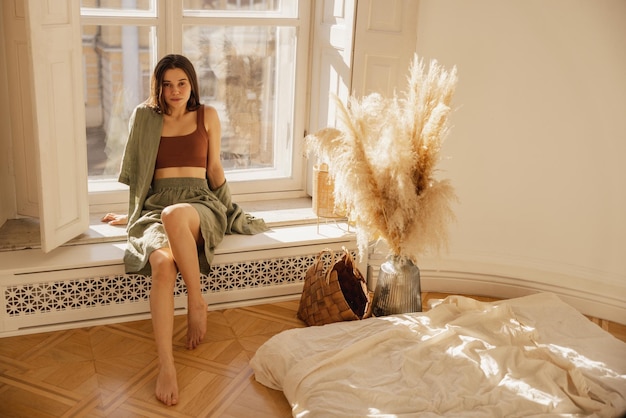 Slim caucasian young girl looks at camera while sitting on windowsill by window in interior of room Brunette wears top shirt and shorts in summer Relax leisure at home indoors