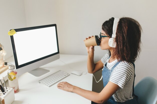 Slim curly brunette girl wears striped shirt typing on keyboard and looking at screen with interest