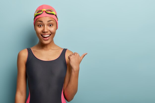 Slim dark skinned female swimmer in black bathingsuit, points thumb on free space
