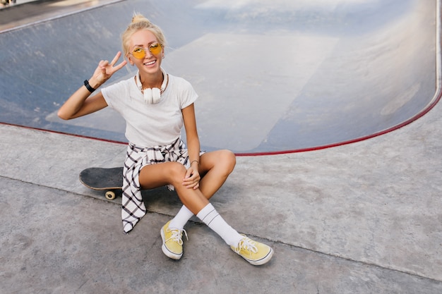 Slim enthusiastic woman in white socks sitting on skateboard. Outdoor photo of glad caucasian woman in white t-shirt having fun in skate park.