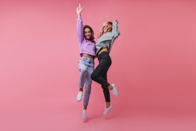 Slim fashionable ladies enjoying life. Magnificent female friends jumping and waving hands.