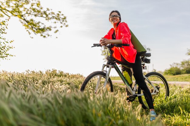 Стройная красивая женщина занимается спортом утром в парке, катается на велосипеде с ковриком для йоги в красочной фитнес-экипировке, исследует природу, улыбается, счастливый здоровый образ жизни