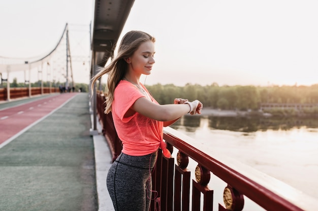 Slim girl in gray pants standing outdoor and looking at smartwatch. Cheerful european lady posing on the street with fitness bracelet.