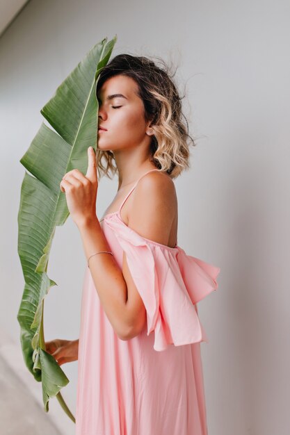 Slim girl in long pink gown posing with eyes closed and holding leaf. Indoor photo of thoughtful young woman with wavy hairstyle sniffing flower.