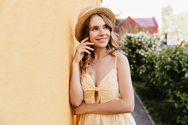 Slim girl in trendy checkered dress standing on the street. Outdoor portrait of good-looking young woman in straw hat.