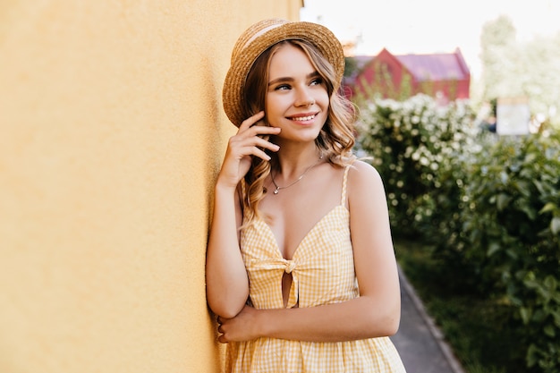 Худенькая девушка в модном клетчатом платье стоит на улице. Открытый портрет красивой молодой женщины в соломенной шляпе.