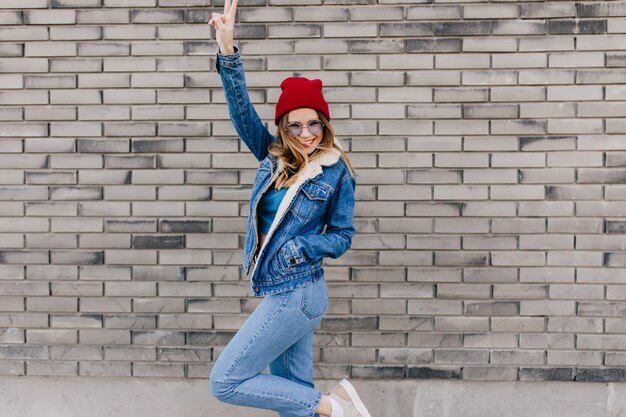 Худенькая девушка в модных джинсах развлекается на улице в холодный весенний день. Блаженная женская модель в джинсовой одежде танцует на городской стене и машет руками.