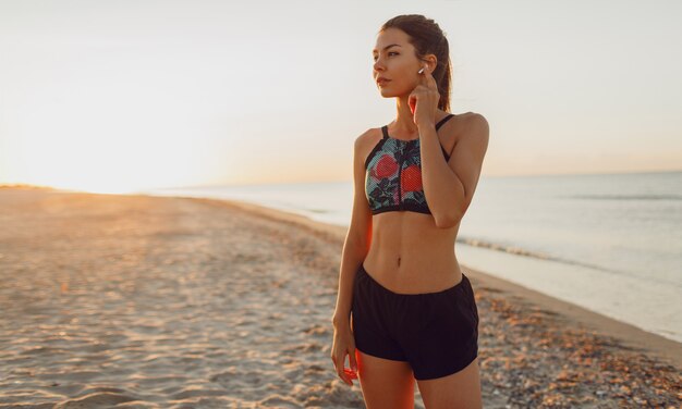 Slim graceful female standing on the beach and relaxing after intense workout.Brunette woman listening music taking break after exercising outdoors.
