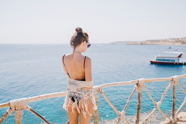 海の景色を楽しみ、打ち上げを見ているトレンディなヘアスタイルのスリムで優雅な女の子。海の上に立っている黒い水着で素晴らしい若い女性の後ろからの肖像画
