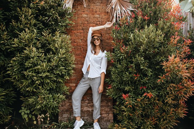 Slim lady in white sneakers, gray pants and white oversized blouse poses against brick wall surrounded by bushes.