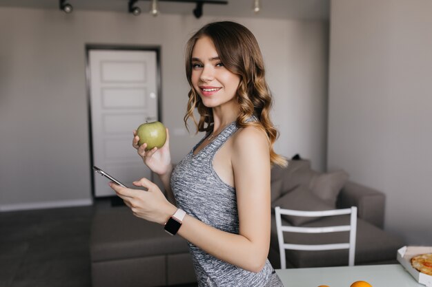 Худенькая бледная девушка в smartwatch позирует дома. Очаровательная темноволосая дама с яблоком веселится утром.