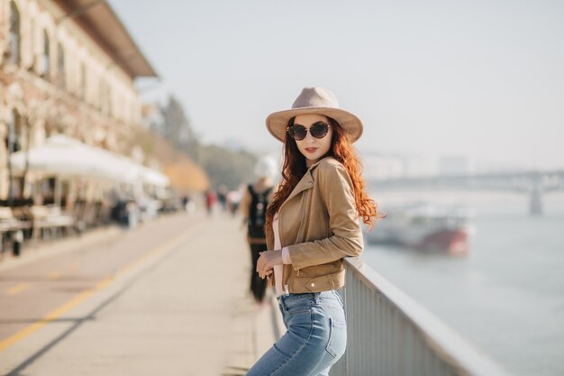 Slim red-haired woman in trendy hat standing in confident pose near sea