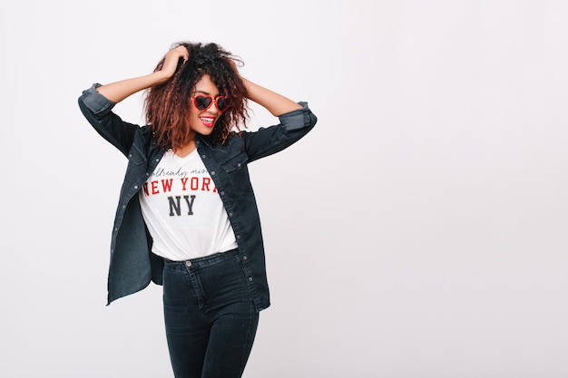 Free photo slim smiling girl wears jeans and sunglasses dancing in studio enjoying photoshoot. young african female model playing with her curly hair and laughing on white background.