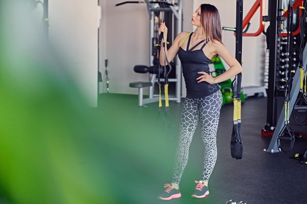 Slim sporty female posing near trx straps stand in a gym club.