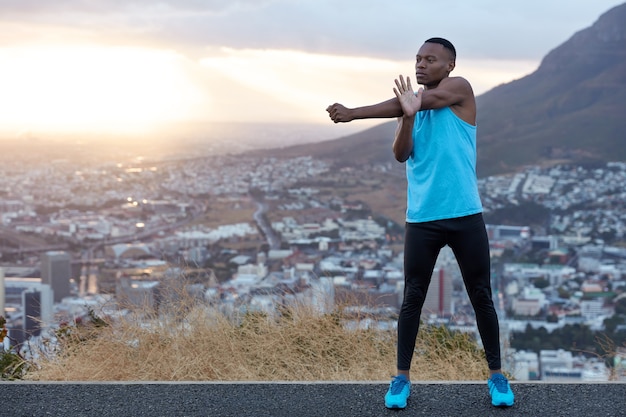 Slim sporty man with strong body, does stretching exercises for hands, prepares for morning run, stands behind mountainous landscape with free space for your promotional content. Sport concept