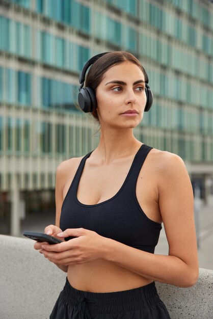 Slim sporty woman looks away pensively uses mobile phone for chatting listens music via headphones dressed in active wear has regular morning training in urban setting. People and sport concept