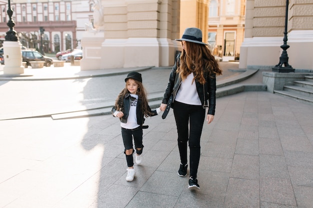 Slim stylish woman in black jeans holding hand of her cute daughter in leather jacket, walking down the street.