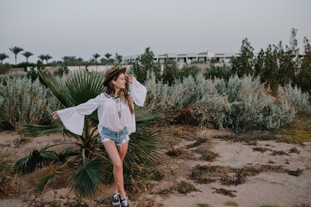 Slim stylish woman in white blouse and denim shorts walking outside enjoys views of exotic plants and beautiful sky. Attractive young woman in sportive shoes gladly posing with dreamy face expression