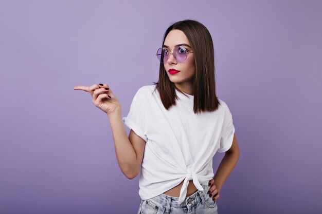 立っているスリムな驚きの女の子。驚いてポーズをとる白いTシャツを着たエレガントな女性の屋内写真。