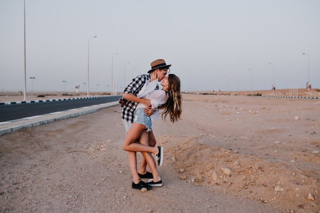片足でデニムのショートパンツに立っているスリムな女性が、美しい砂漠でボーイフレンドに優しくキスします。夏休みに高速道路に近いポーズ彼のガールフレンドを抱きしめるスタイリッシュな若い男