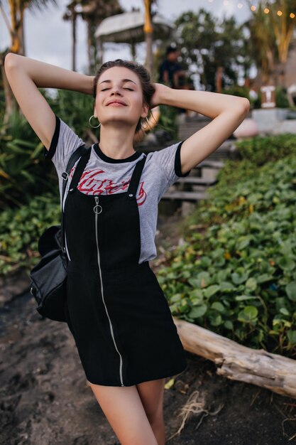 Slim woman in vintage casual attire posing with eyes closed. Outdoor portrait of graceful young lady in trendy t-shirt enjoying life in summer at resort.
