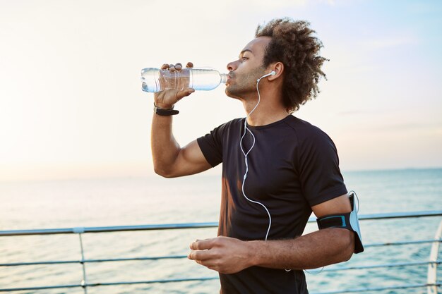 Стройный и молодой спортсмен освежается, пьет воду из пластиковой бутылки в лучах утреннего солнца. Слушал свои любимые песни во время пробежки.