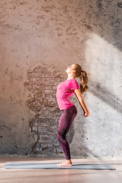 Slim young woman practicing stretching exercise standing against weathered wall