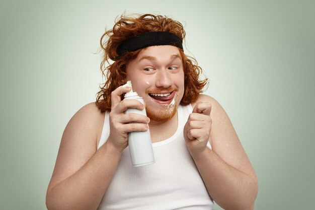 Sly redhead fat male in sports band and tank top holding spray can, having crazy joyful look