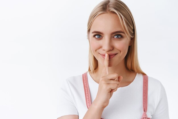 Sly and sassy attractive feminine blond woman with blue eyes, smiling secretly telling you rumors, press finger to lips seal with promise untell anyone, standing white background shushing