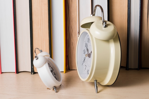 Small alarm clock in front of large clock in front of bookshelf on wooden desk