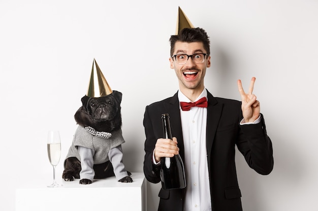 Small black dog wearing party hat and standing near happy man celebrating holiday, owner showing peace sign and holding champagne bottle, white background.
