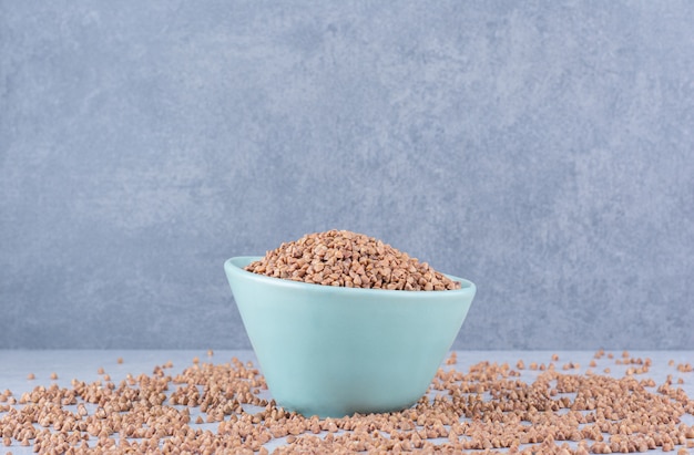 Small bowl of buckwheat placed in the middle of scattered grains on marble surface