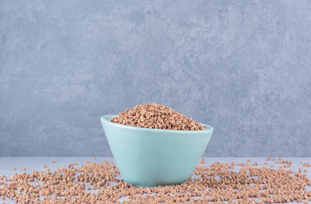 Small bowl of buckwheat placed in the middle of scattered grains on marble surface