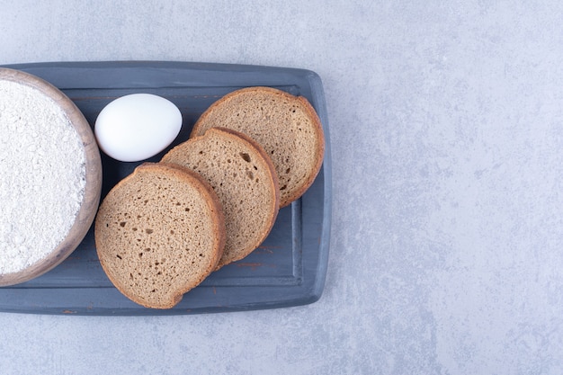 Small bowl of flour, an egg and three bread slices on a navy board on marble surface