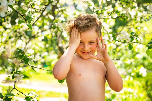 A small boy in a flowery garden holds his hands to his head and smiles, standing with his bare torso