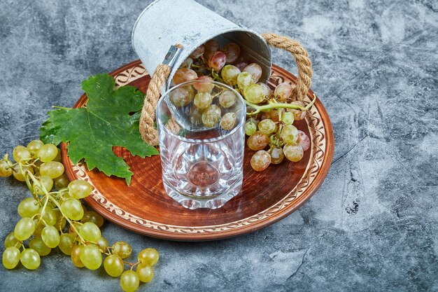 Небольшое ведро с виноградом внутри керамической тарелки и стакана на мраморном фоне. Фото высокого качества