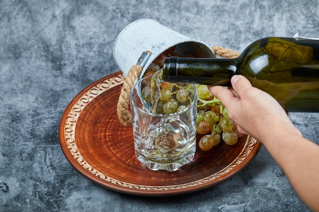 Small bucket of grapes inside ceramic plate and hand pouring wine into the glass on marble.