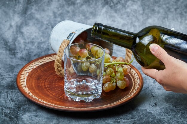 Small bucket of grapes inside ceramic plate and hand pouring wine into the glass on marble.