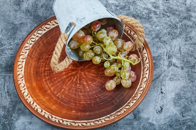 Небольшое ведро с виноградом внутри керамической тарелки на мраморе.