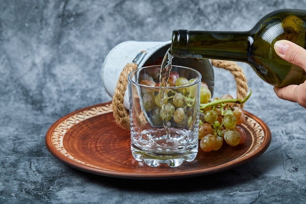 Бесплатное фото Небольшое ведро с виноградом внутри керамической тарелки и рука наливает вино в бокал на мраморном фоне