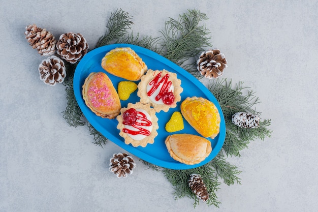 Small buns, cupcakes and jelly candies on a blue platter decorated with pine leaves and cones on marble surface