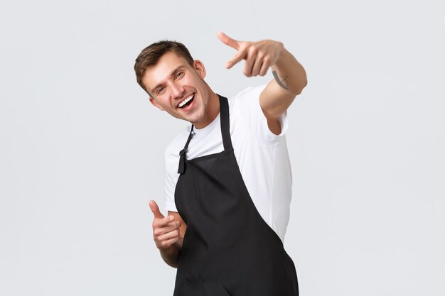 Small business owners, coffee shop and staff concept. Friendly handsome barista dancing and pointing finger at camera, inviting you to enjoy drinks in cafe, smiling happy, choosing guest