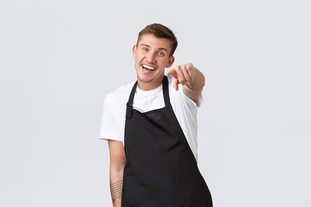 Small business owners, coffee shop and staff concept. Friendly handsome barista, waiter in apron pointing at camera, inviting you to visit cafe and enjoy drinks, delicious dessert, white background