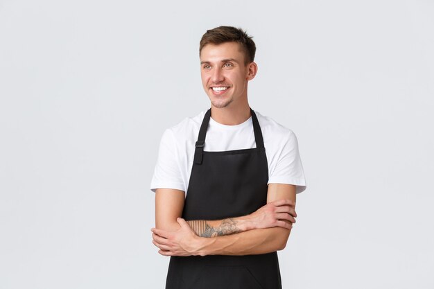 Small business owners, coffee shop and staff concept. Handsome cheerful blond guy barista, part-time worker looking pleased upper left corner with satisfied smile, wearing apron