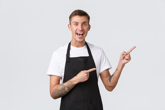 Small business owners, coffee shop and staff concept. Happy good-looking part-time worker in cafe, barista or waiter in black apron pointing fingers upper right corner and smiling upbeat