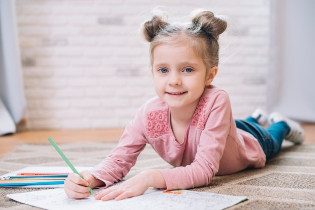 Small cute girl drawing in book lying on carpet at home