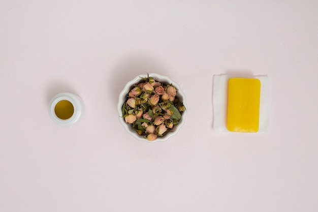 Маленькие сухие бутоны роз в керамической белой миске; бутылка меда и травяное желтое мыло на салфетке на текстурированном фоне