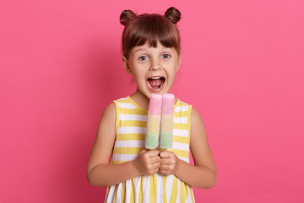 Small European kid biting fruit ice cream, little charming girl with widely opened mouth, wearing summer clothes, looks happy, having fun with delicious dessert.