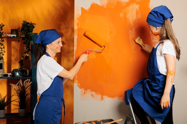 Free photo small family using orange paint on walls, young child on ladder painting surface with paintbrush and mother using roller brush tool. people having fun with housework renovation, home decor.